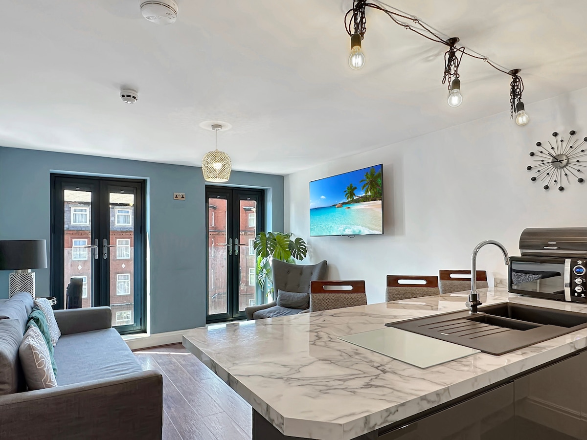 A modern kitchen area features a marble countertop and sleek appliances. A comfortable seating arrangement is visible, with large windows providing ample natural light. A television displaying a serene beach scene enhances the inviting ambiance of the open space.