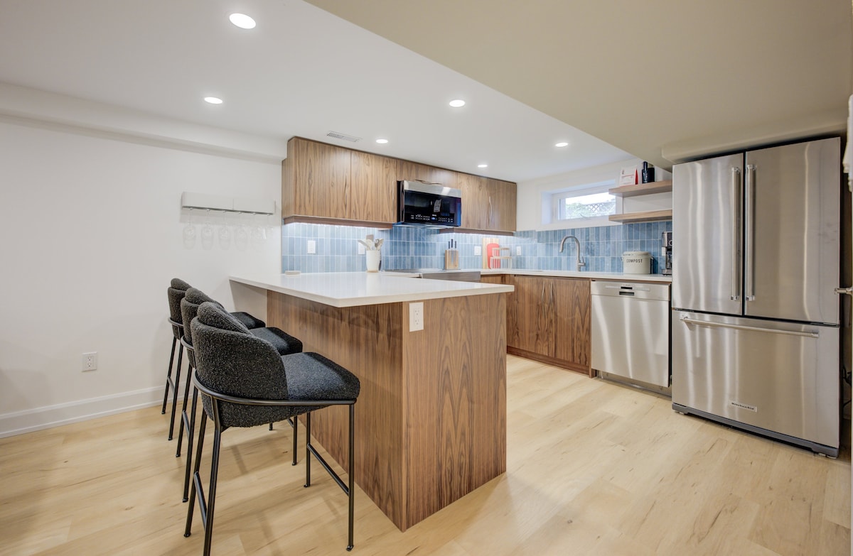 A modern kitchen space is highlighted by sleek wood cabinetry and a large island with seating for four. Stainless steel appliances are visible, including a refrigerator and dishwasher. Soft lighting creates a bright atmosphere, while a blue tile backsplash adds a touch of color.