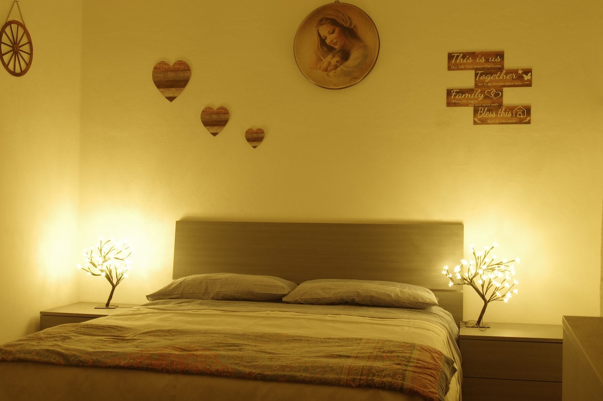 A calm bedroom features a spacious bed with soft linens, complemented by warm lighting from decorative lamps on either side. Heart-shaped decor and a wall hanging add personal touches, while a serene image enhances the tranquil atmosphere of the space.