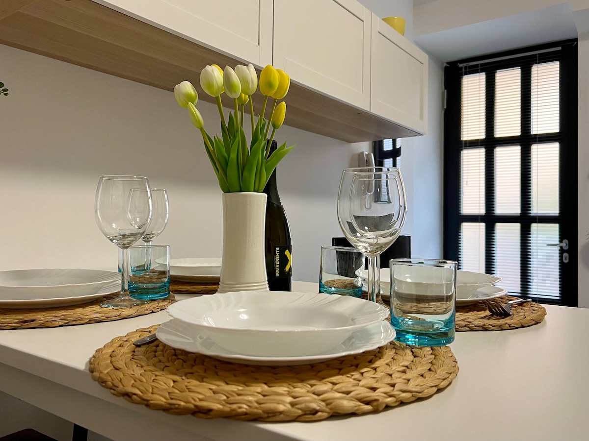A dining table is set with white plates on woven placemats, accompanied by glasses for wine and water. A vase with yellow tulips adds a touch of greenery, while a bottle is positioned nearby. Sunlight filters in through a window with black frames.