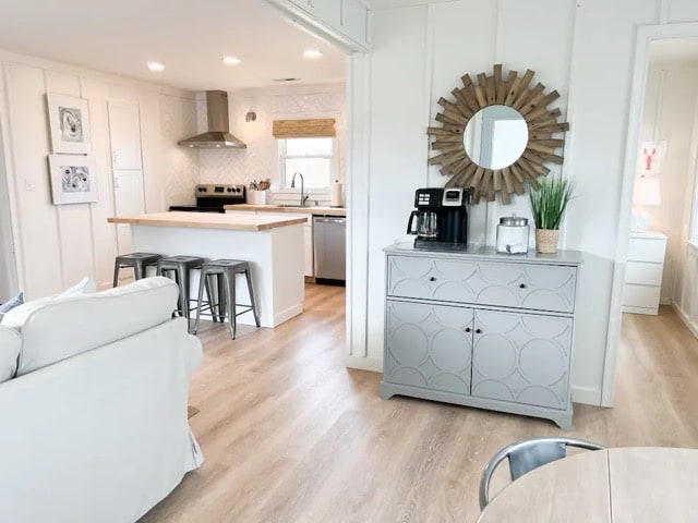 An open-concept living space showcases a modern kitchen with stainless steel appliances and a wooden island. Light-colored cabinetry and decor create a bright atmosphere. A round dining table and gray console table are visible, accompanied by a decorative mirror and greenery, enhancing the welcoming environment.