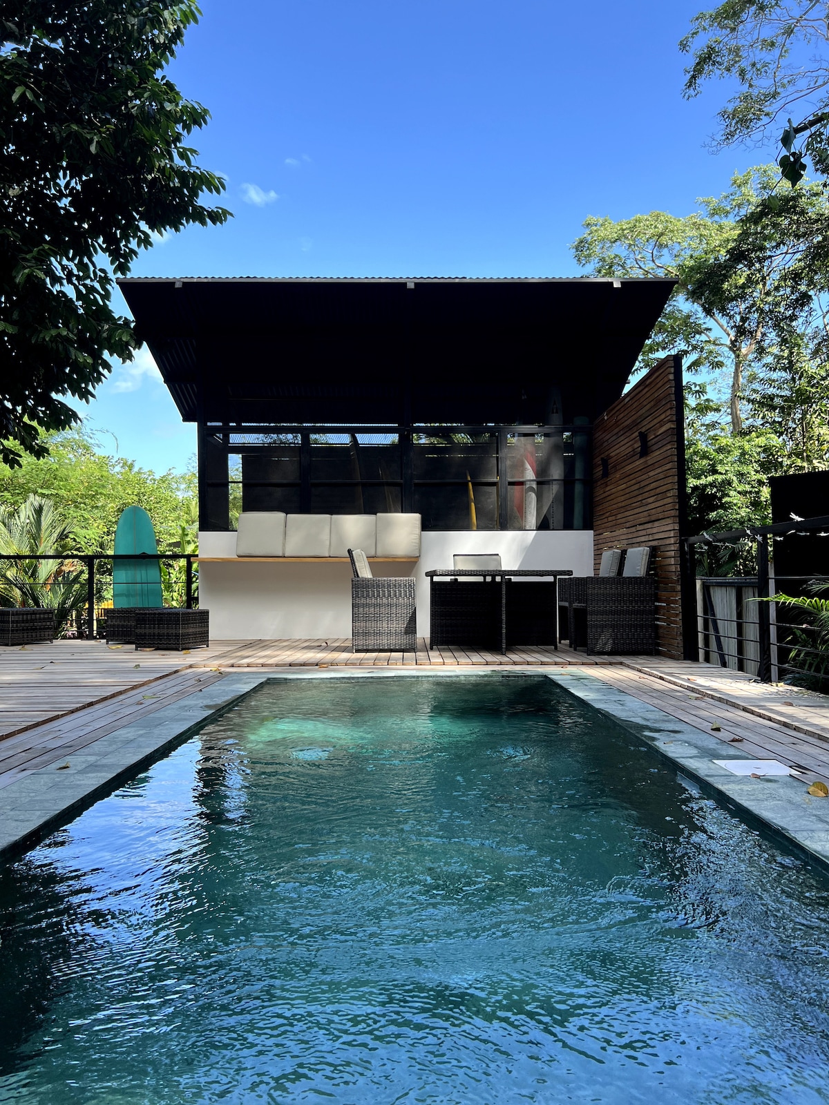 A modern casita is visible featuring large glass doors that open to a wooden deck and a sparkling pool. Comfortable seating arrangements are placed on the deck, with lush greenery surrounding the area, creating a serene outdoor setting.