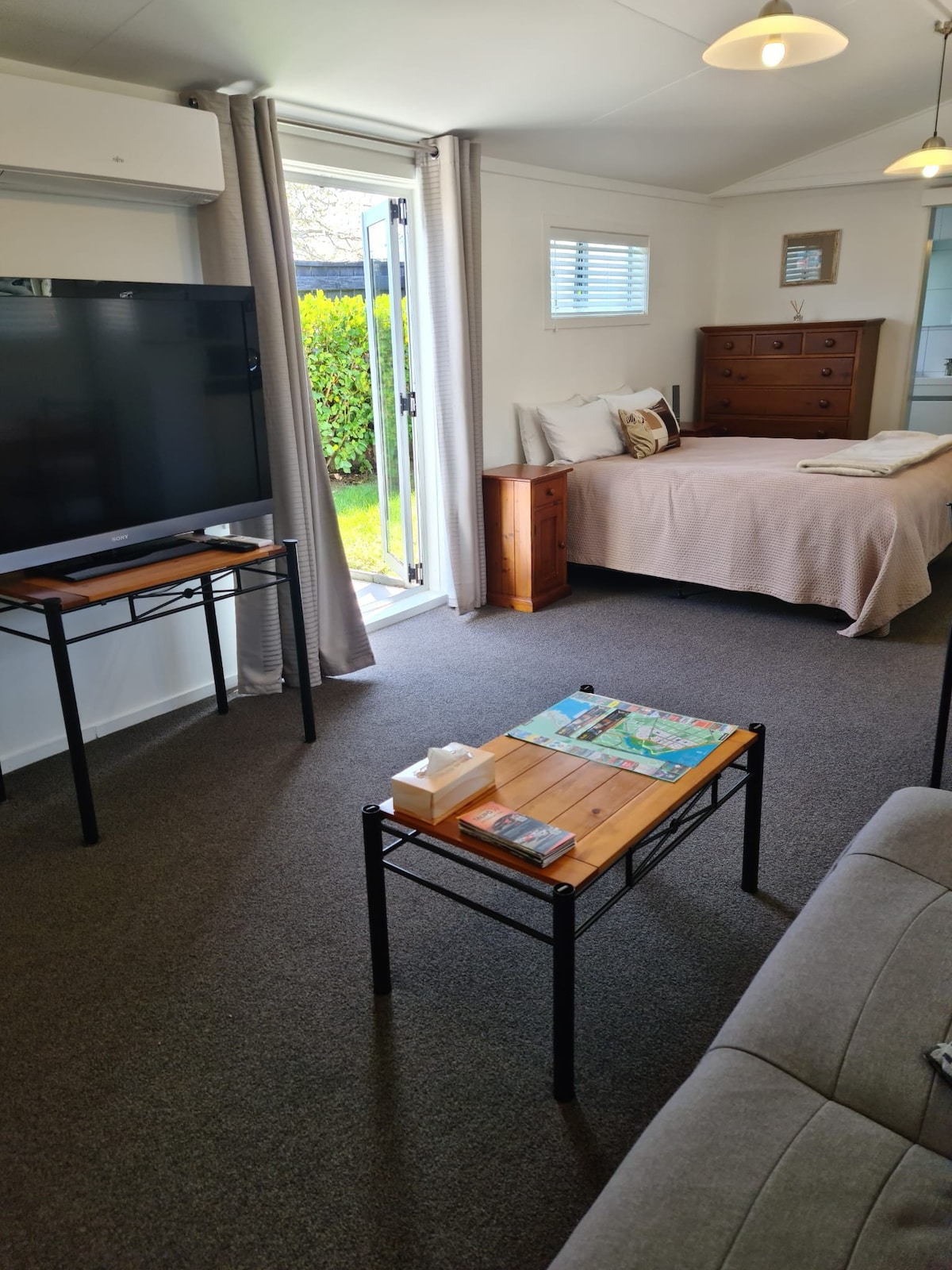 A spacious studio is presented with a sitting area featuring a comfortable couch and a wooden coffee table displaying a map. A large television is positioned against the wall, and a cozy sleeping area with a neutral-toned bed can be seen. Light enters from the open door leading to an outdoor space.