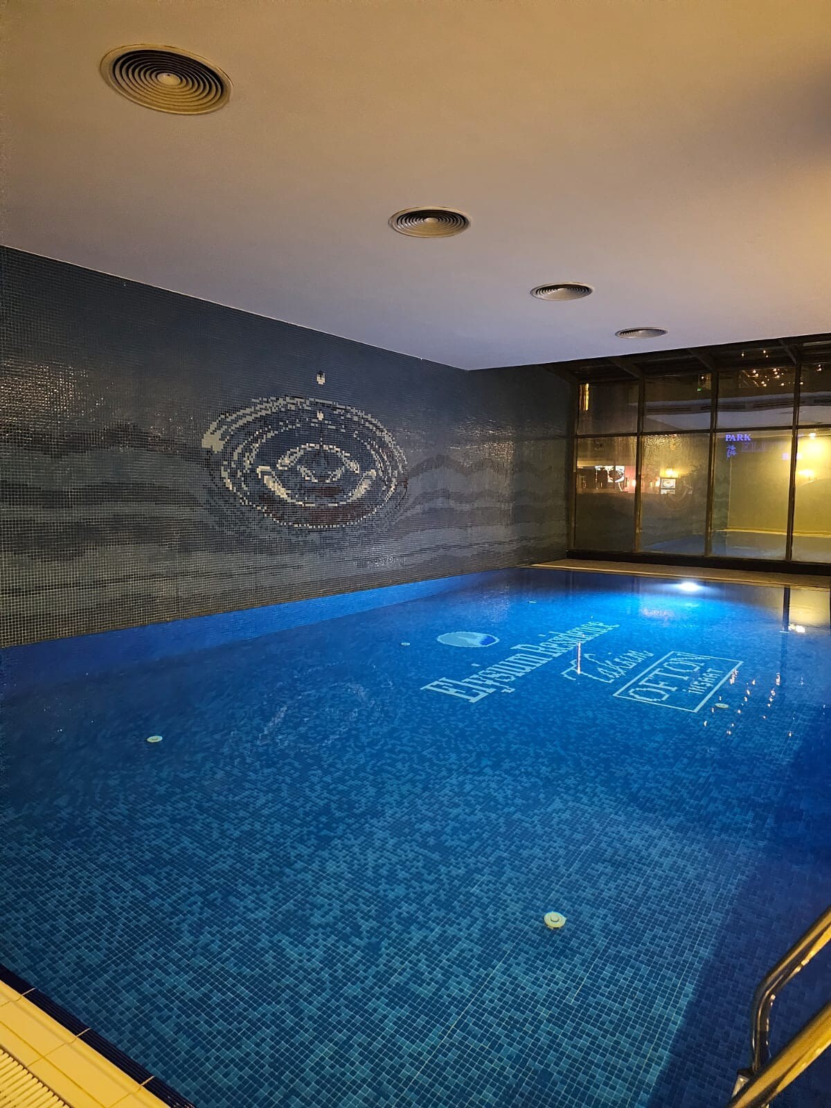 An indoor pool is illuminated with soft blue lighting, highlighting the tiles' intricate patterns. A large mural can be seen on the far wall, while windows allow for a glimpse of the evening exterior. The atmosphere conveys a serene and private space for relaxation.