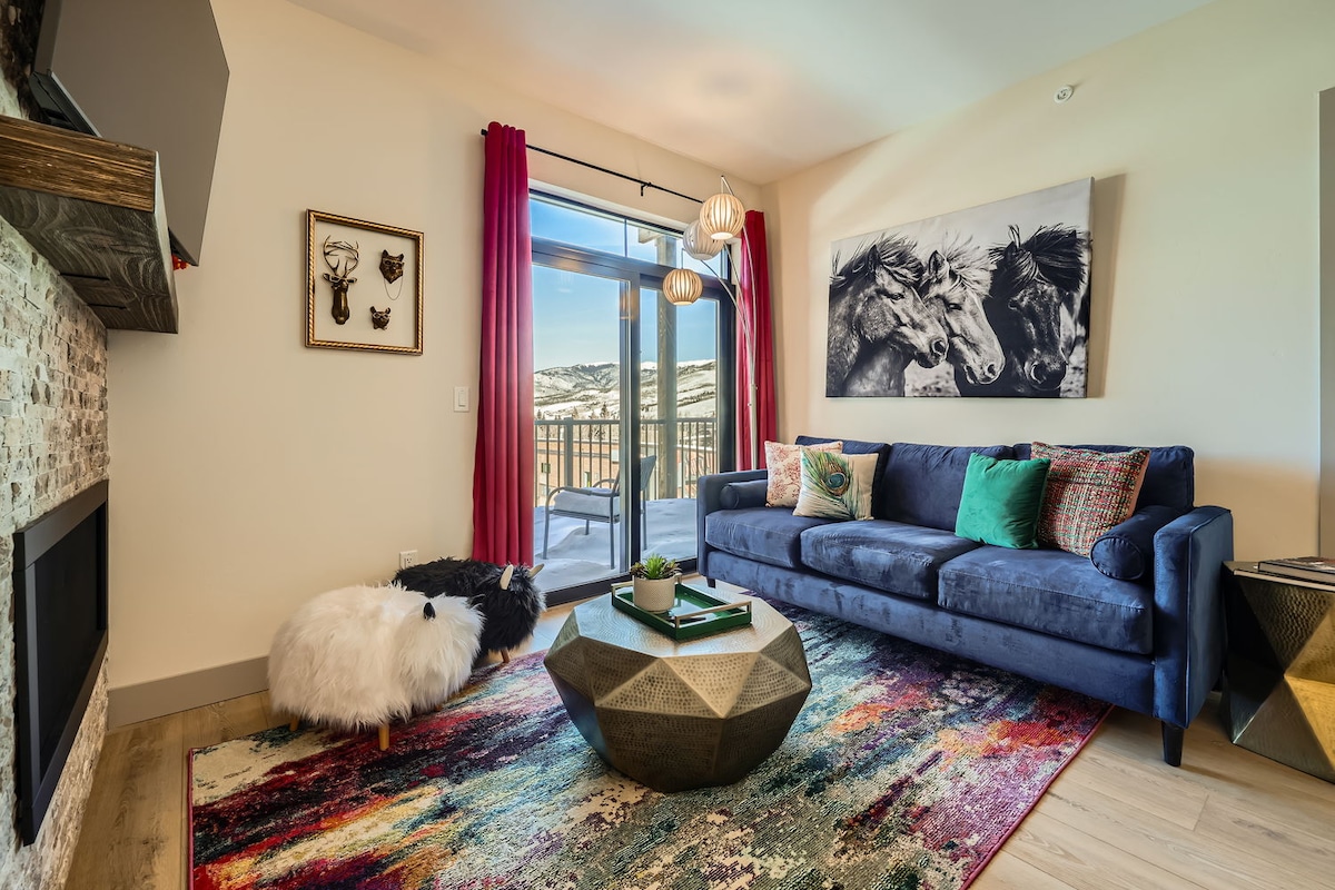 A modern living area features a navy sofa adorned with colorful throw pillows, complemented by a geometric coffee table. A vibrant area rug adds warmth to the wood flooring. Large windows provide natural light and access to a balcony, while a stone fireplace serves as a focal point.