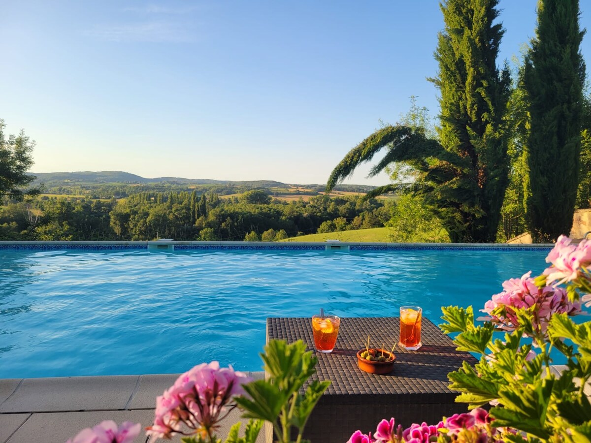 A serene pool area is surrounded by vibrant flowers with a scenic view of rolling hills in the background. A table is set with two drinks and a small bowl of snacks, inviting relaxation and enjoyment of the tranquil landscape.