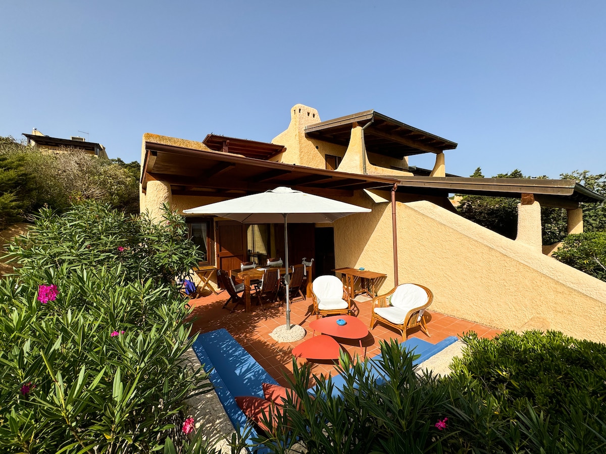 The exterior of the two-story beach house is adorned with soft yellow walls and features a spacious patio area. Comfortable seating arrangements are under a large umbrella, surrounded by lush greenery and colorful flowers, creating a serene outdoor space for relaxation.