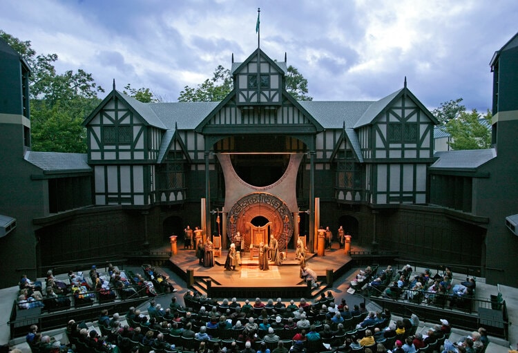 An open-air theater is depicted, showcasing a stage adorned with intricate scenery. Rows of audience seating are filled with guests watching a performance, surrounded by lush trees. The building reflects a traditional architectural style, with detailed wooden beams and a close-knit, inviting atmosphere.