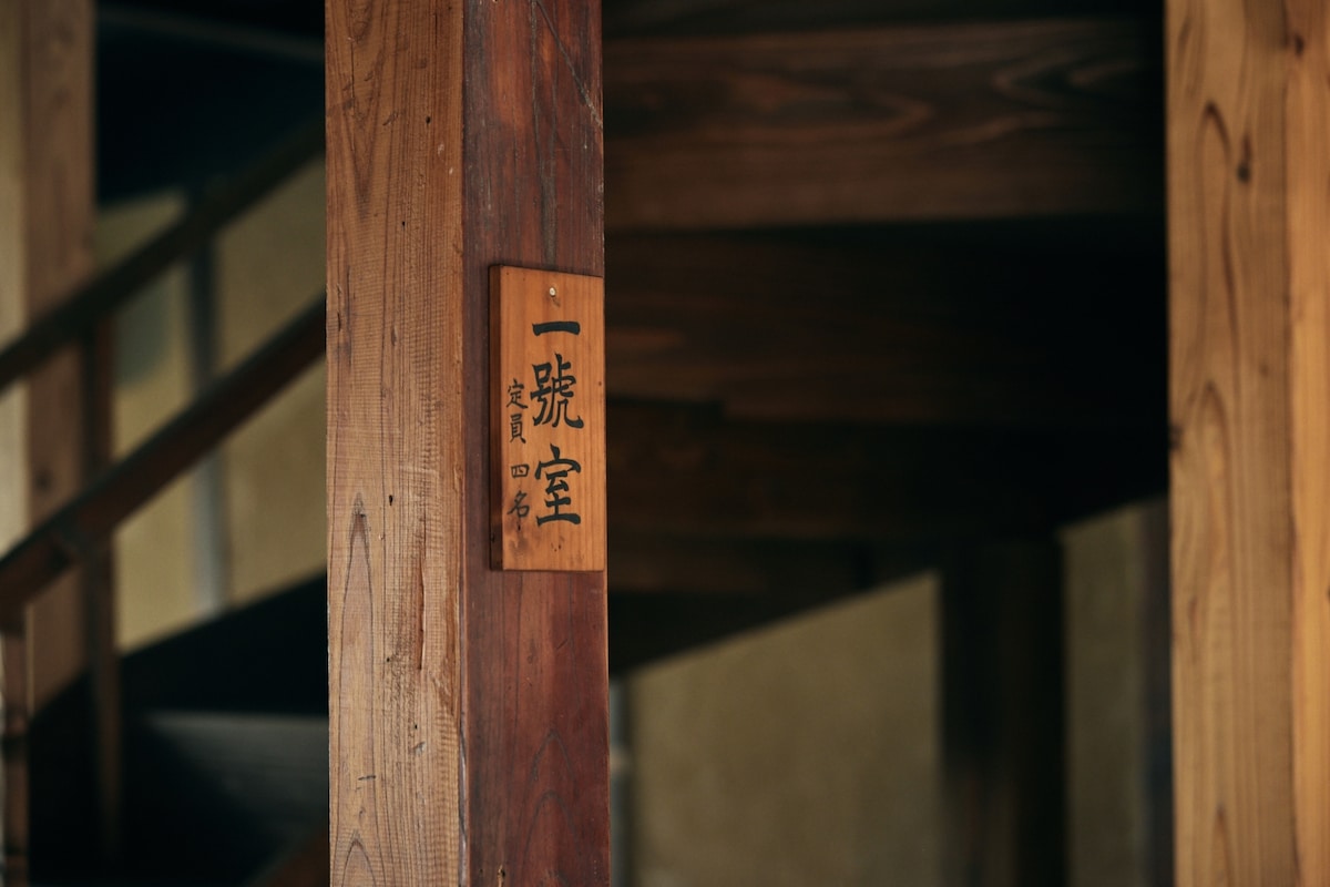 A wooden post features a sign marked with the characters for 'Room 1.' The post is part of a staircase, highlighting the natural wooden texture of the structure. The soft lighting creates a warm ambiance in the hallway.