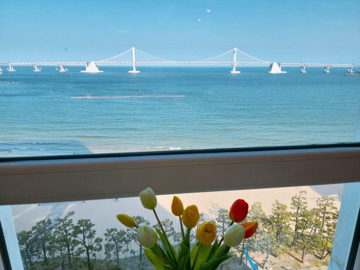A view from the window reveals the expansive ocean and the iconic bridge in the distance. Clear blue skies reflect over the water, while a vase of colorful tulips sits in the foreground, adding a touch of vibrancy to the serene landscape.