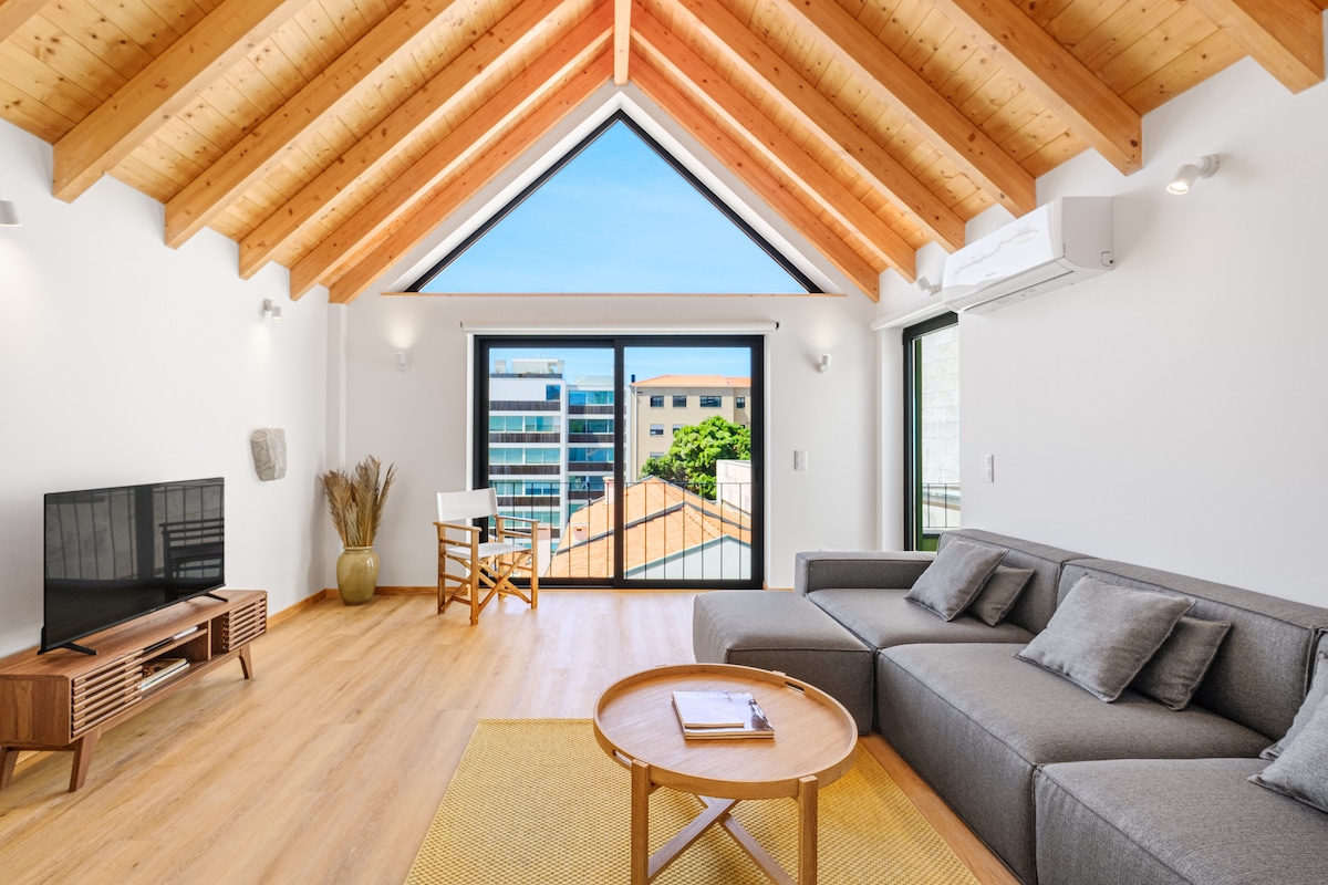 The living area features high wooden ceilings with exposed beams, allowing ample natural light. A comfortable gray sectional sofa is complemented by a circular wooden coffee table, while a TV is positioned on a low profile media console. Large glass doors open to an outdoor view.