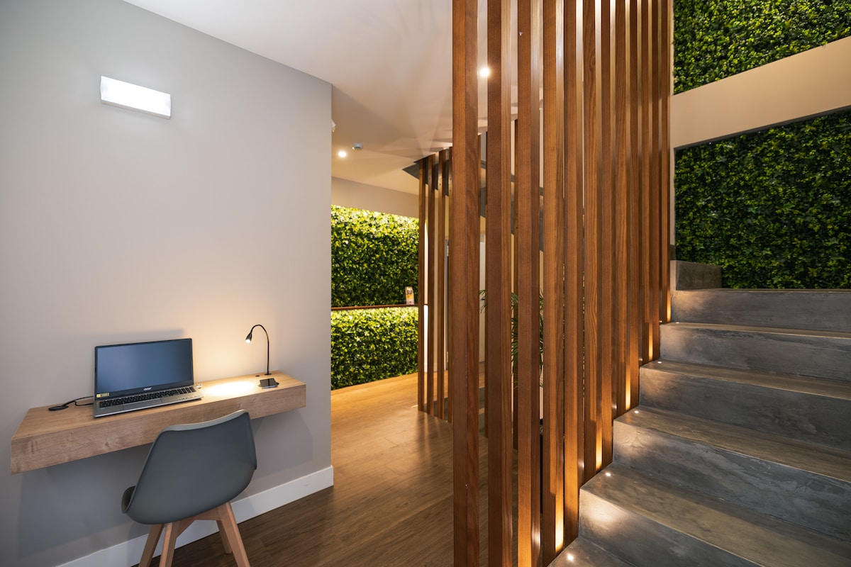 A modern entryway features a wooden desk with a laptop and a sleek lamp. The space is enhanced by vertical wooden slats and a wall of green plants, while a staircase leads to the upper level, illuminated by soft lighting.