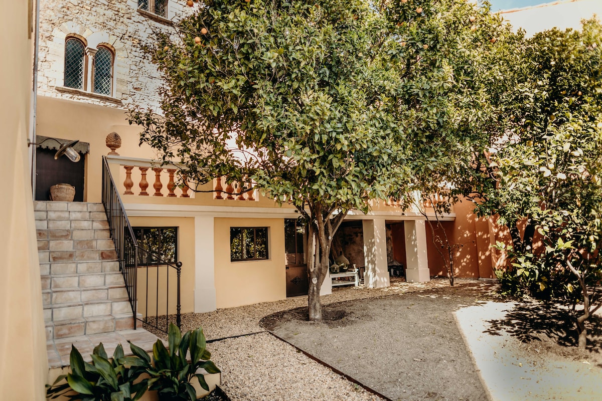A serene courtyard is surrounded by lush greenery and a large citrus tree, providing a sense of tranquility. A stone staircase leads to the upper level of the property, while the well-maintained ground features light-colored gravel, enhancing the inviting atmosphere.