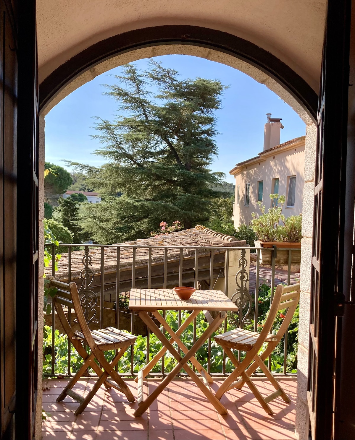 A small balcony is framed by an archway, featuring a wooden table and two chairs. Lush greenery is visible beyond the railing, with terracotta rooftops and a distant building providing a scenic view.