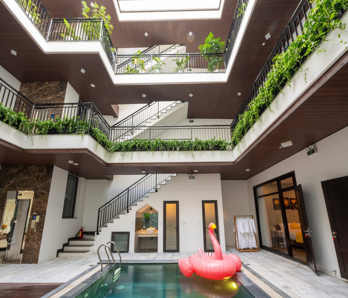 The interior courtyard showcases a modern swimming pool surrounded by lush greenery and multiple levels of balconies. A pink inflatable flamingo floats in the pool, adding a playful touch. Elegant staircases lead to upper floors, enhancing the villa's spacious layout.