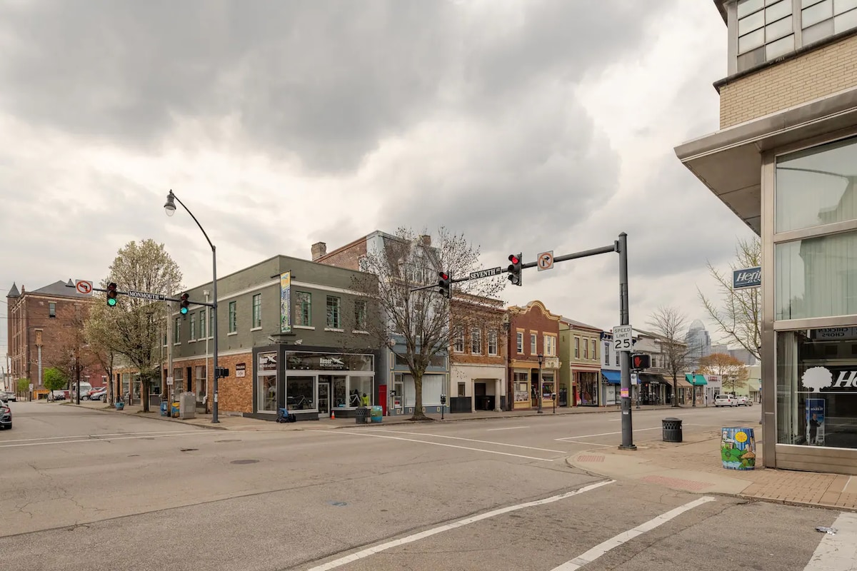 A street view showcases a quiet intersection in a vibrant downtown area. Various buildings, including retail and commercial spaces, line the street. Traffic lights and street signage are visible, while a few trees add greenery to the scene. The overcast sky creates a subdued atmosphere.