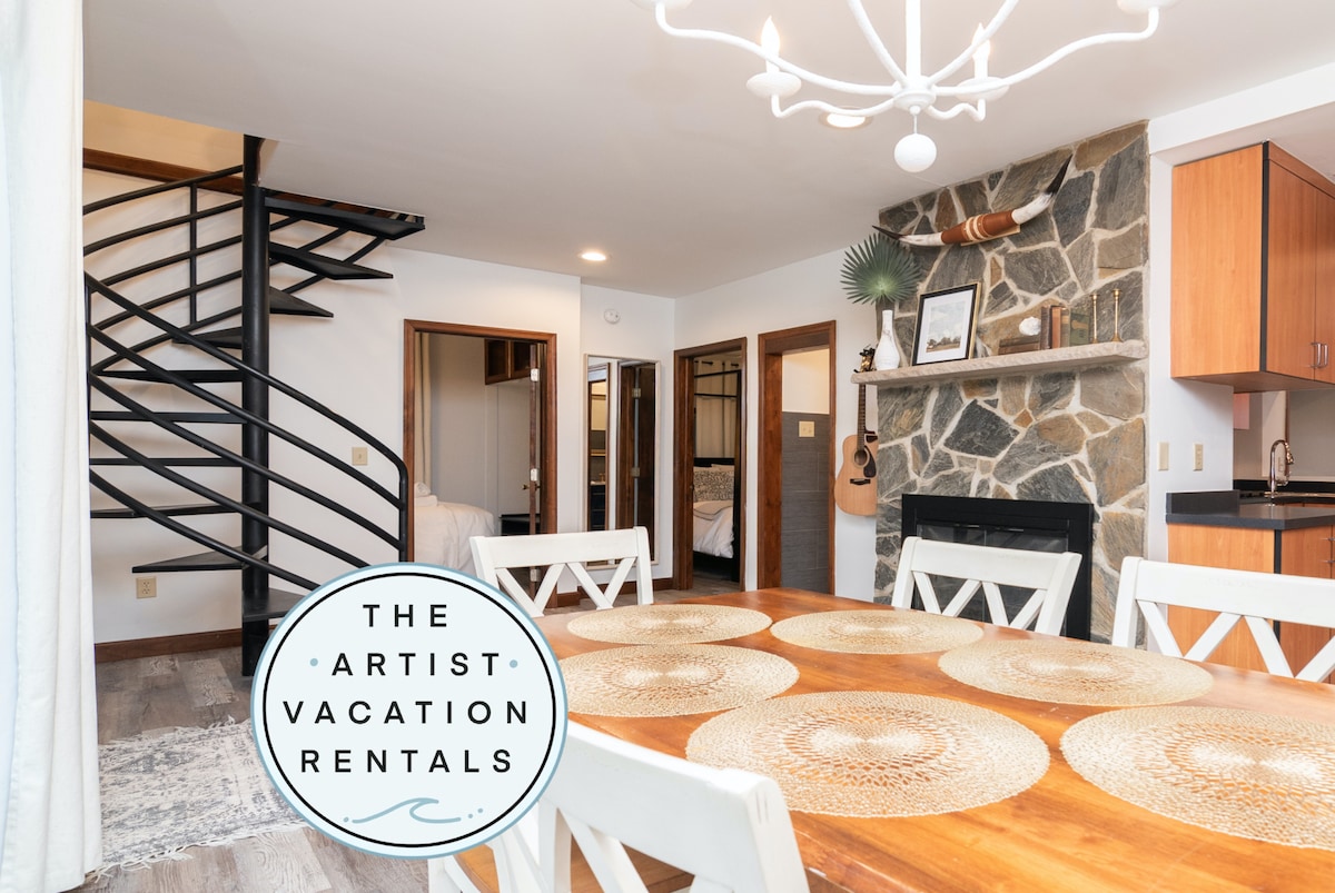 A spacious dining area is highlighted by a large wooden table surrounded by white chairs. A unique spiral staircase is visible in the background, leading to the upper level. A stone fireplace adds warmth to the room, while modern kitchen cabinetry is seen on the right.