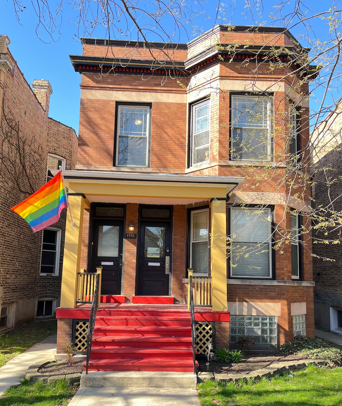 The two-story brick building features a welcoming entrance with a red staircase leading up to the front door. A rainbow pride flag is prominently displayed, symbolizing inclusivity. Large windows allow natural light to brighten the exterior, surrounded by well-maintained greenery.