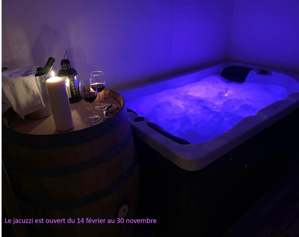 A private jacuzzi is illuminated with soft blue lighting, creating a calming atmosphere. Nearby, a candle stands next to a wine glass and a bottle of wine, set atop a wooden barrel, inviting relaxation and enjoyment.