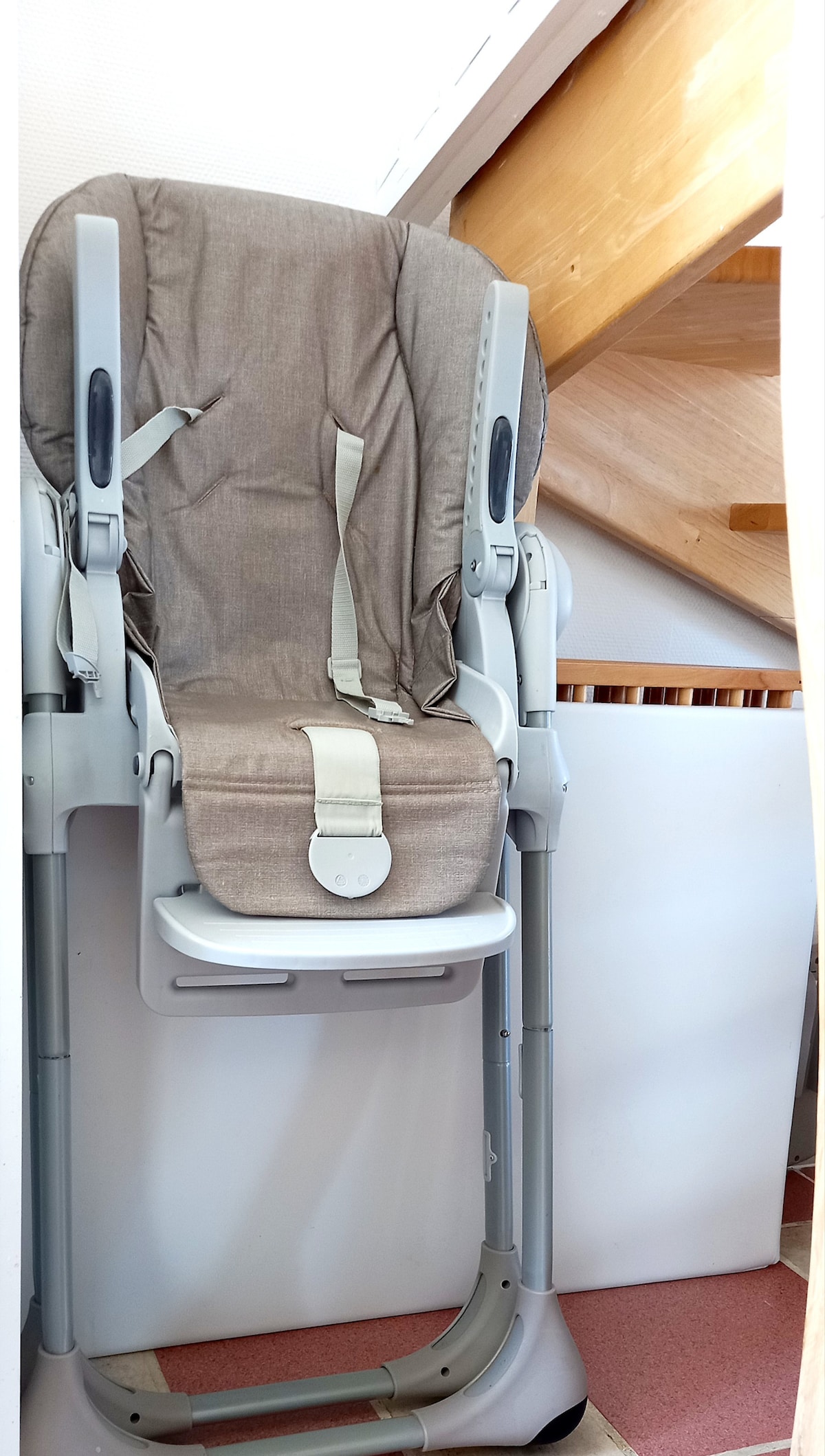 A high chair is positioned next to a staircase, featuring a neutral-toned padded seat and safety straps. The structure is designed for child safety with adjustable height options, ensuring easy access for caregivers.