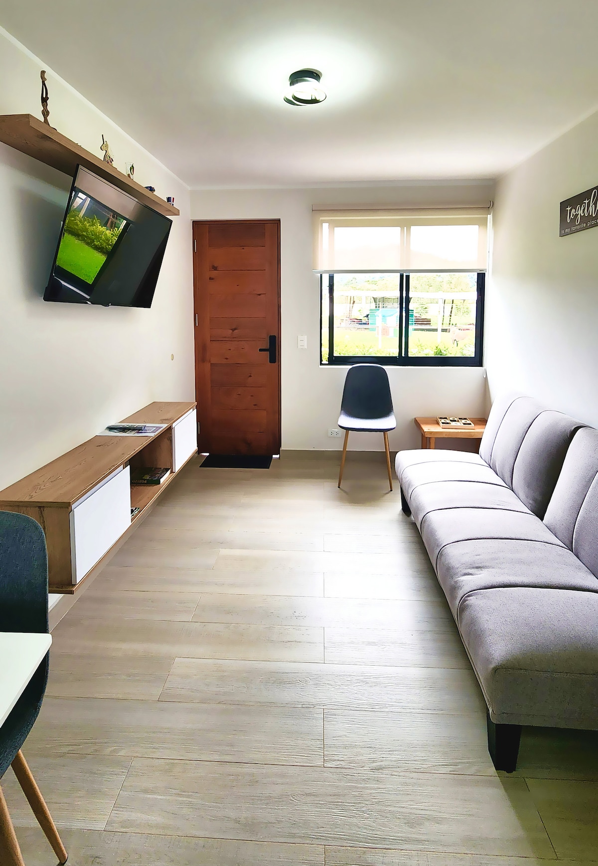 A bright living area features a grey futon, providing comfortable seating for relaxation. A wall-mounted television is positioned on a wooden shelf, and natural light enters through the window, which is set with two black frames. The space is complemented by a modern chair and simple decor.
