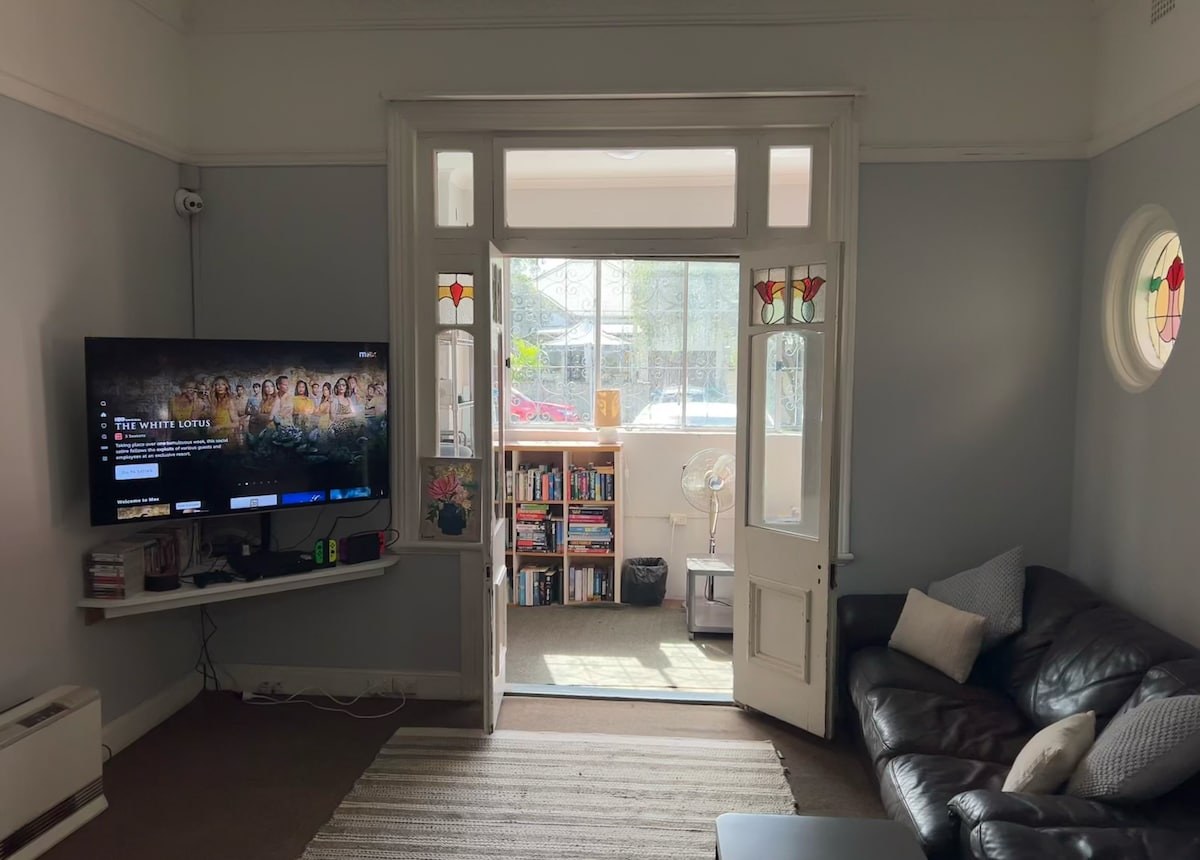 A comfortable living area features a dark leather sofa positioned near a wall-mounted television. Shelves filled with books are visible in the background, and natural light filters through decorative glass doors leading outside.