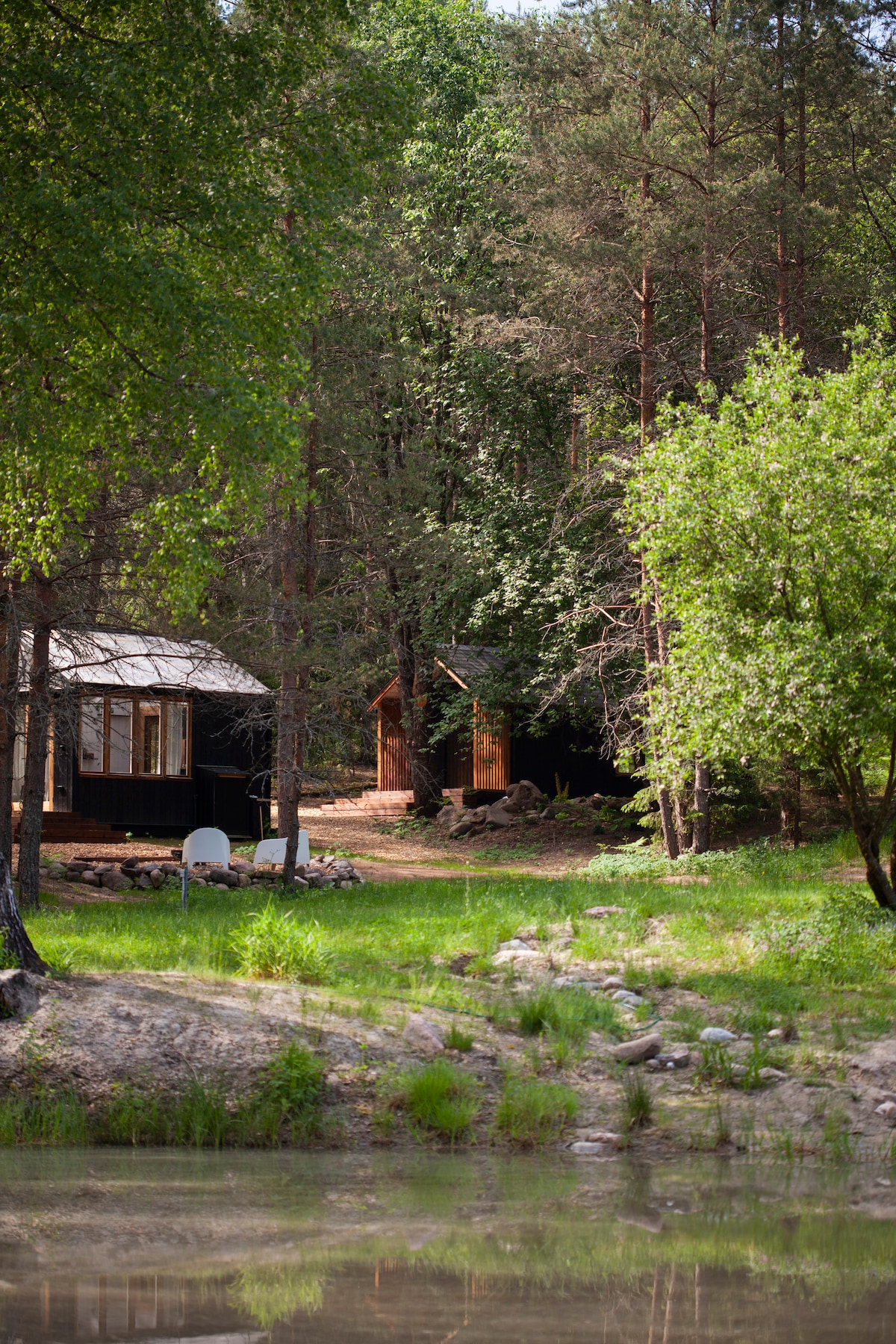 A serene outdoor setting is depicted, featuring two wooden cabins nestled among lush greenery and trees. A pond reflects the surroundings, while grassy areas provide a natural landscape leading to the cabins. The peaceful atmosphere invites relaxation and connection with nature.