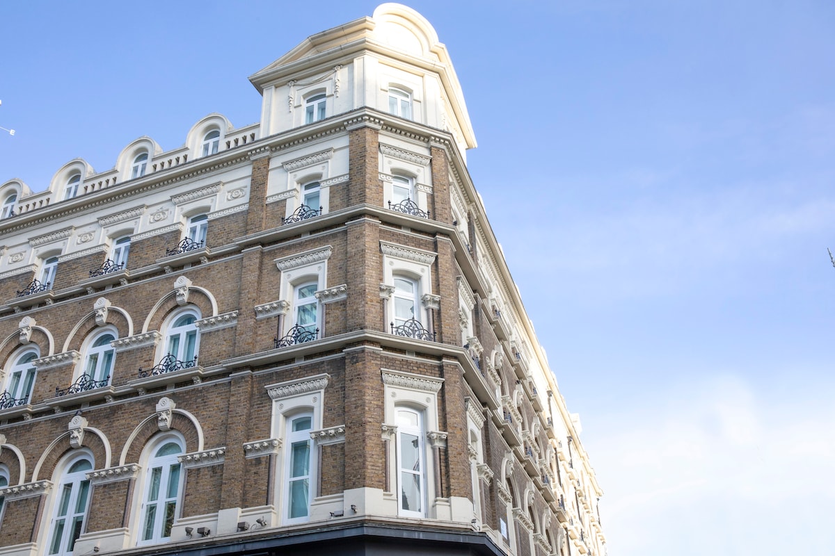 The exterior of a historic building showcases a classic Georgian façade. The brick structure features ornate detailing, multiple windows, and decorative balconies, with a peaked roof accentuating its height. Clear blue skies provide a contrasting backdrop to the architectural elegance.