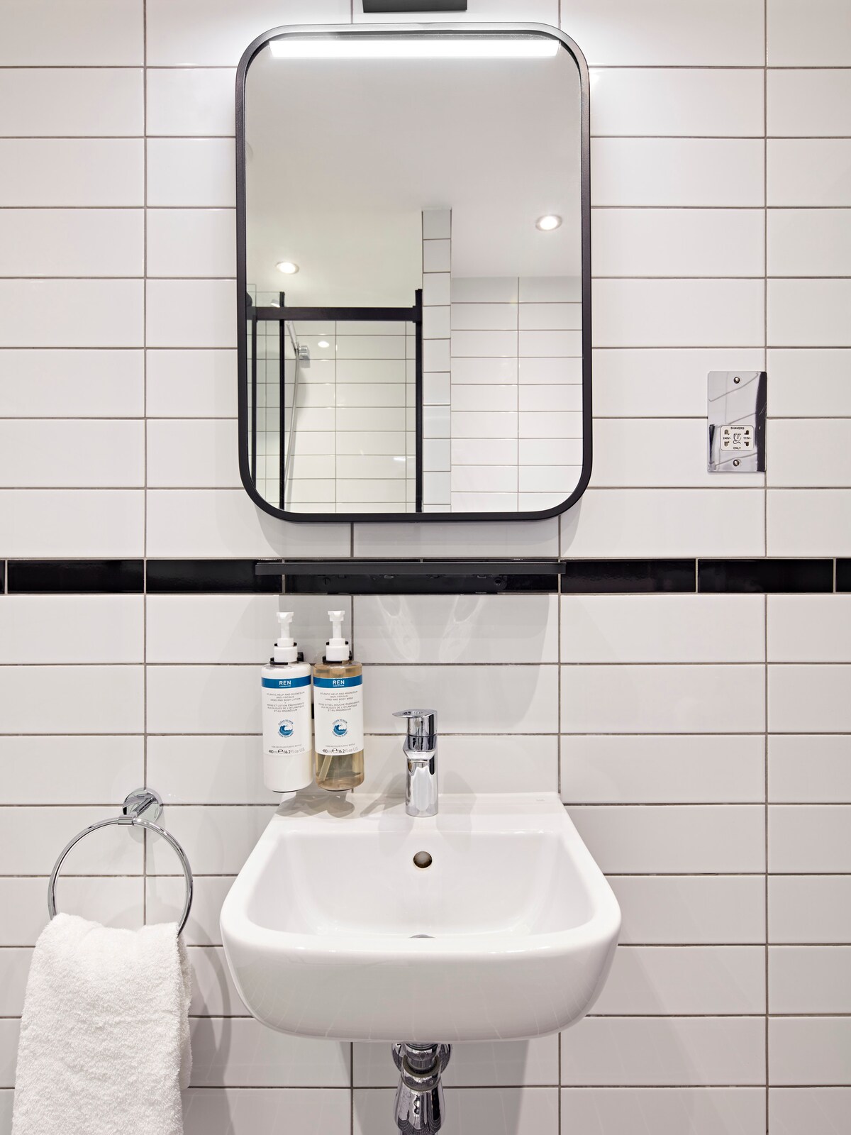 A modern bathroom features white tiled walls with a contrasting black line. A compact sink is displayed below a rounded mirror, accompanied by wall-mounted dispensers for toiletries. A towel hangs neatly from a nearby silver ring, enhancing the clean and organized ambiance.