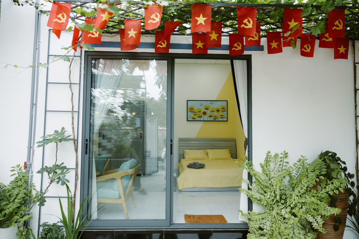 An inviting exterior view showcases a glass sliding door leading into a bedroom. The balcony is adorned with red flags featuring a star symbol, while potted plants add greenery. A cozy bed is visible inside, complemented by a television mounted on the wall.