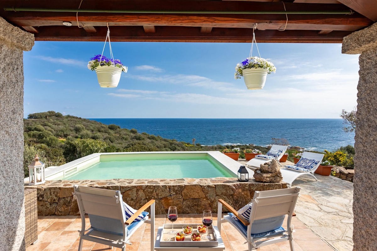 A serene outdoor space features a private pool with a stunning view of the Mediterranean Sea. Two sunbeds are positioned nearby, and a small table holds refreshments, inviting relaxation. Planters with flowers are suspended above, adding a touch of greenery to the scene.