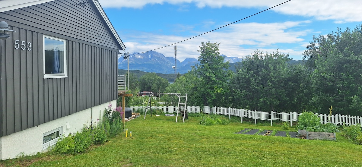The exterior of the apartment is visible, featuring a grassy yard with a swing set and garden plots. A white picket fence surrounds the area. Majestic mountains can be seen in the background under a partly cloudy sky, creating a serene countryside setting.