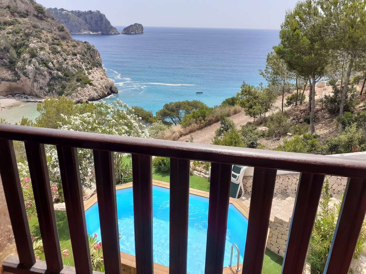 A scenic view reveals a tranquil pool surrounded by lush greenery. The sparkling blue water of the pool contrasts with the expansive sea in the background, while rocky cliffs and coastal vegetation frame the picturesque landscape.