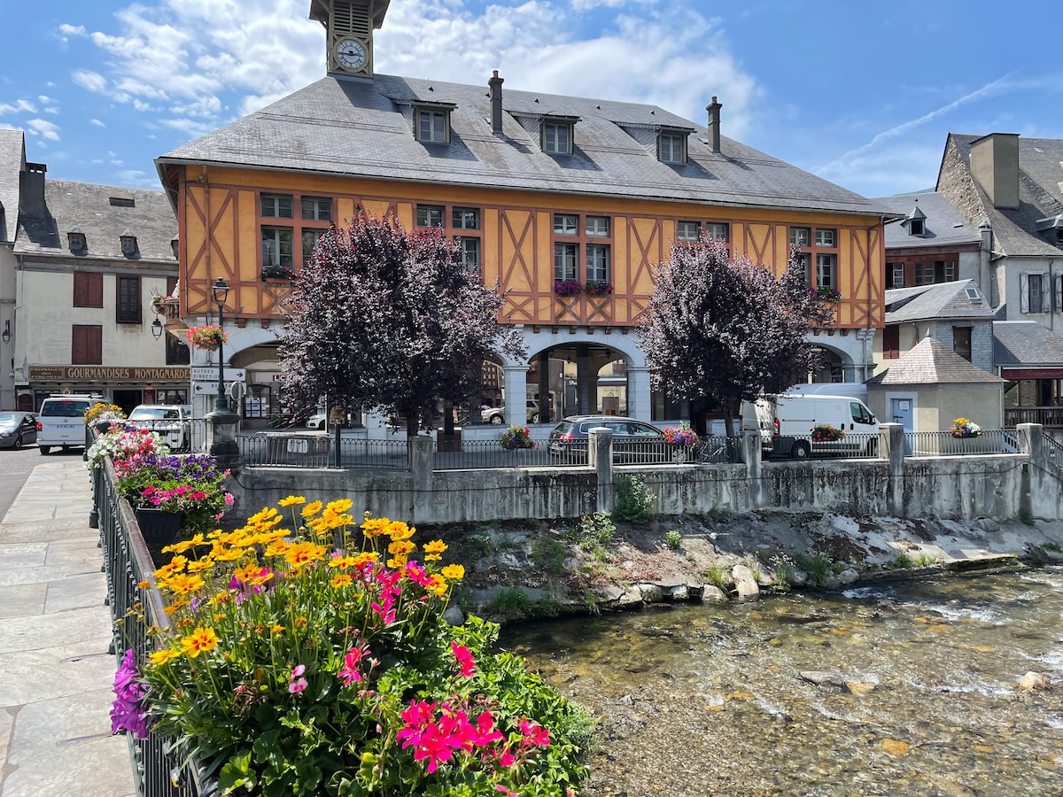 A charming building with a wooden framework features several large windows and is adorned with purple-leaved trees. Colorful flowers in vibrant yellows and pinks line a stone pathway beside a flowing stream, contributing to the scenic view. A clock tower rises above the structure.
