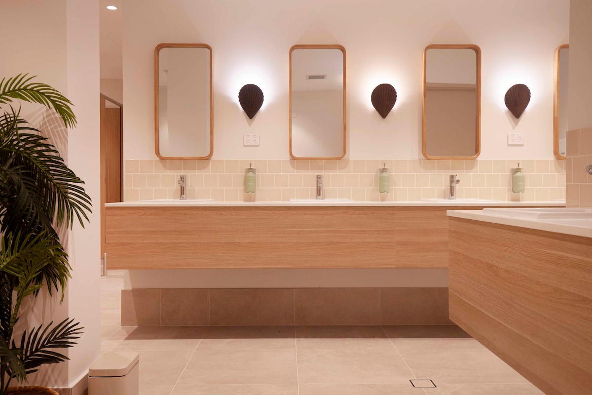 A bright and modern bathroom features four sinks positioned beneath three mirrors, framed in rounded wood. Soft green soap dispensers are placed beside each sink, while stylish wall sconces provide warm lighting. A large potted plant adds a touch of greenery to the space.