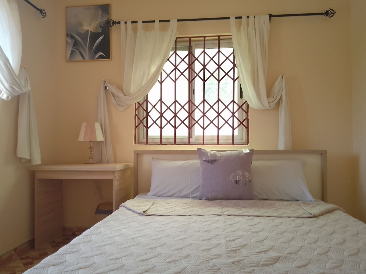 A bright master bedroom features a spacious bed adorned with neutral-toned linens. Natural light enters through a decorative window with a grid pattern, framed by flowing white curtains. A small bedside lamp sits on a wooden desk, enhancing the room's calm ambiance.
