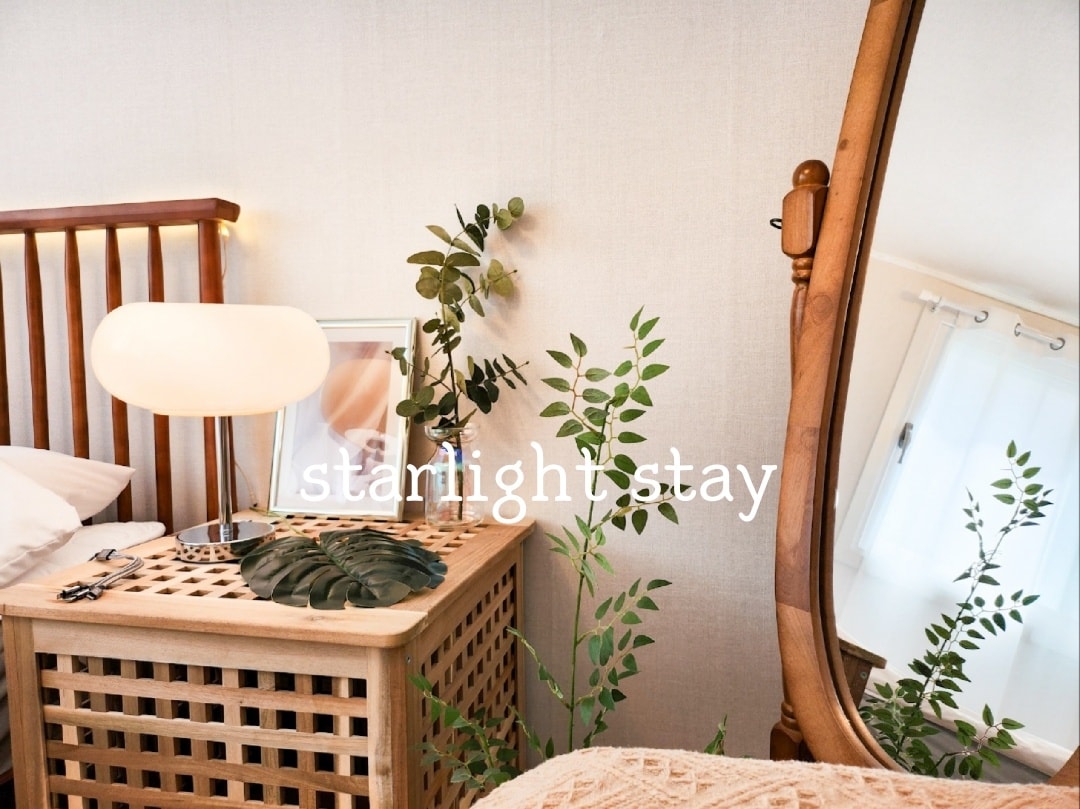 A bedside table crafted from light wood features an elegant lamp and a small mirror. Greenery is displayed on the table, adding a natural touch. A textured blanket drapes over the bed, while a decorative plant is positioned near a window, providing ambiance.