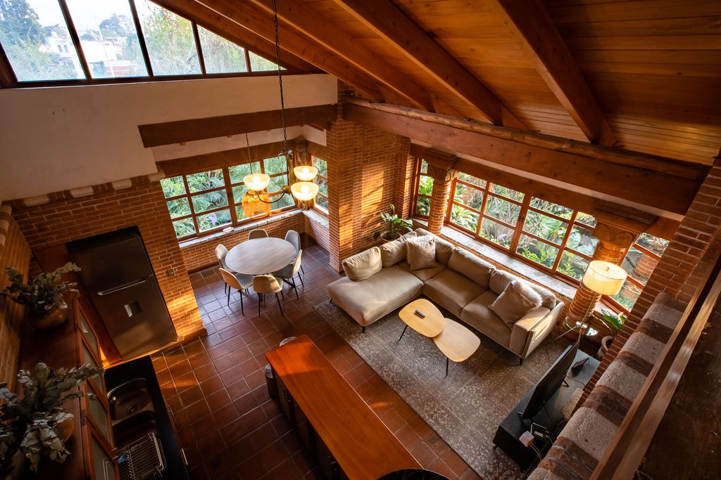 The spacious living area is highlighted by high wooden ceilings and large windows that allow natural light to fill the room. A comfortable sectional sofa and a round dining table complement the brick walls, while a modern coffee table and minimalistic decor enhance the inviting atmosphere.