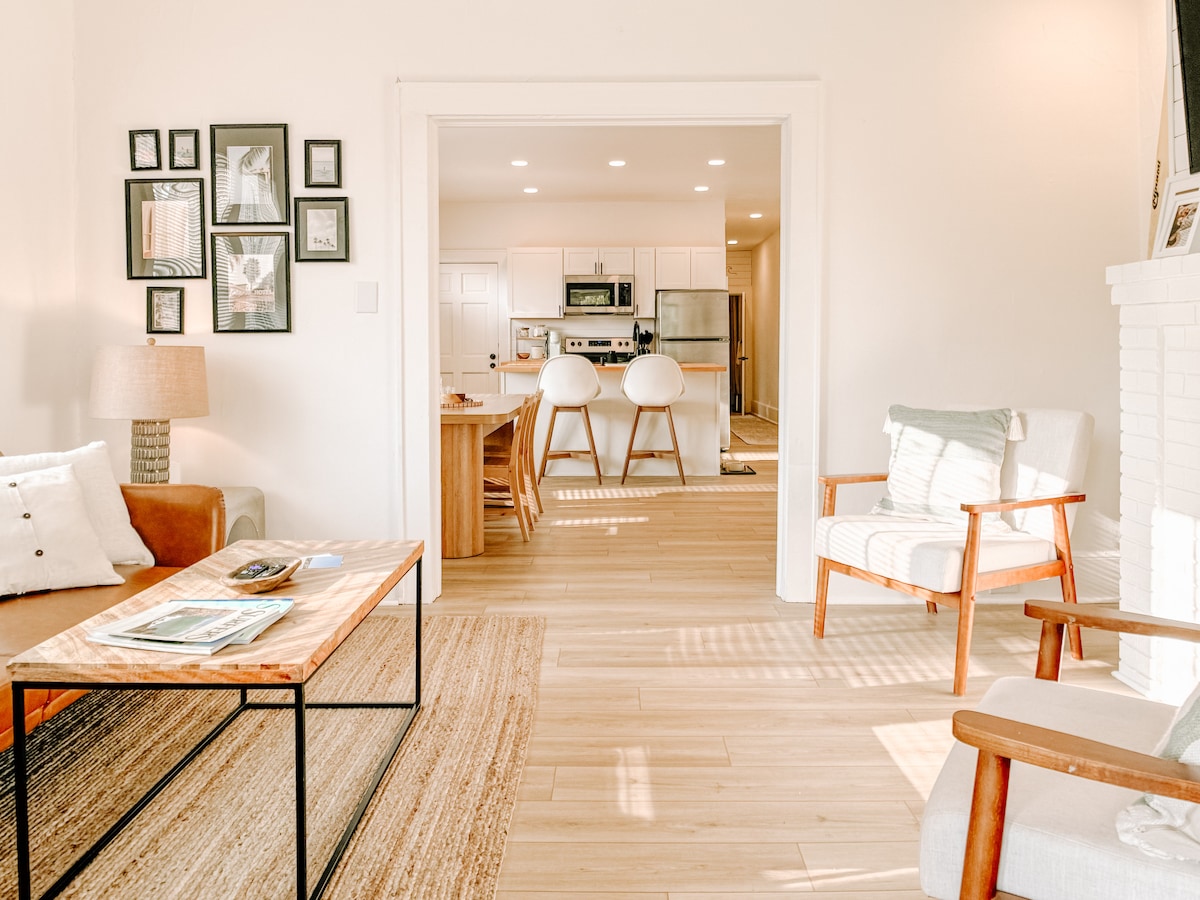 A welcoming living area is visible, featuring a light-colored sofa and a cozy armchair. A wooden coffee table is centered on the floor, with a decorative rug layered beneath. Natural light fills the space, highlighting the open layout that connects to a modern kitchen in the background.