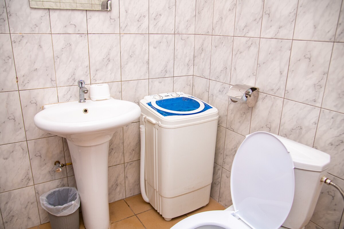 A clean bathroom features a white pedestal sink accompanied by a modern washing machine with a blue top. A toilet is positioned next to the sink, with neutral tile flooring and textured white walls enhancing the space.