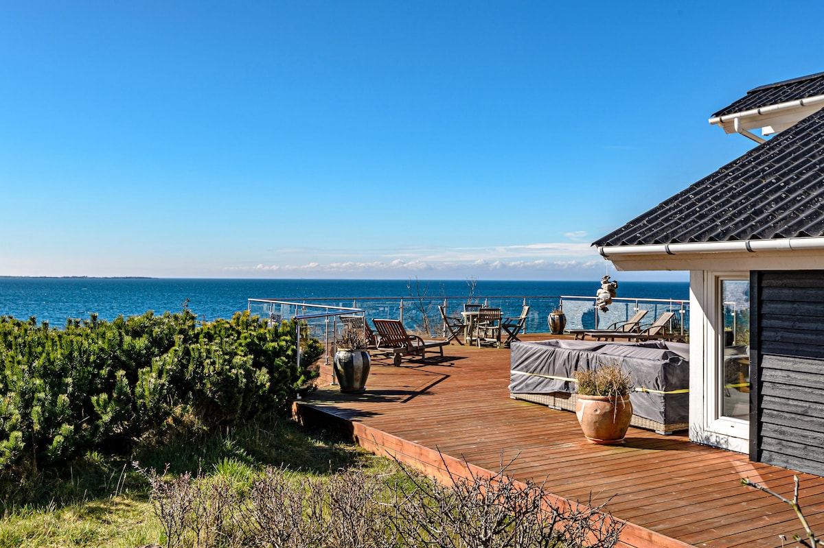 A spacious deck extends from the house, featuring lounge chairs and a hot tub. Planters with greenery are positioned along the edge, while the expansive sea views stretch to the horizon under a clear blue sky.
