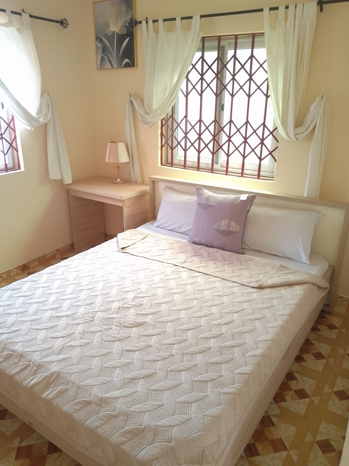 A well-lit bedroom features a spacious bed with a light-colored quilt and a decorative pillow. A bedside table with a lamp is placed next to the bed. Natural light enters through two windows, adorned with sheer drapes. The floor displays a patterned tile design.