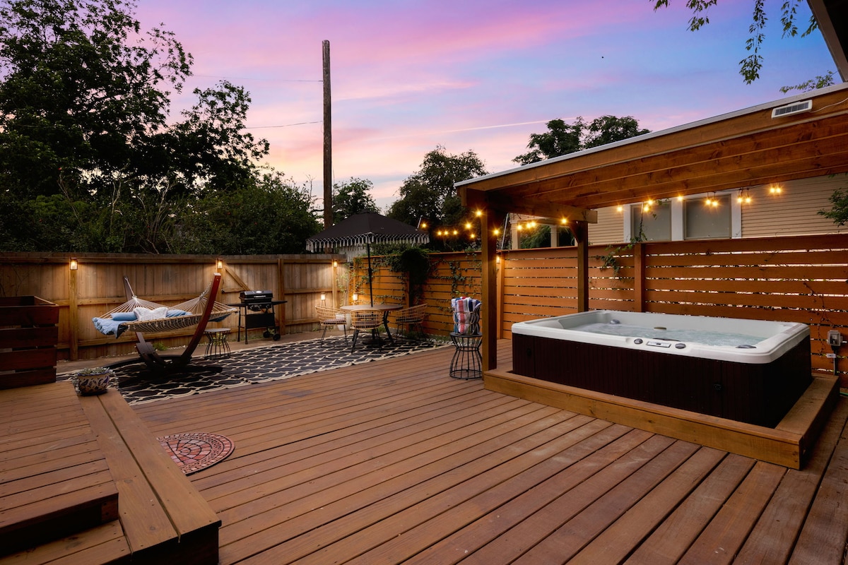 The outdoor patio features a spacious wooden deck illuminated by string lights, creating a relaxing ambiance. A private hot tub occupies one corner, while a hammock and grill complement the space, surrounded by wooden fencing for added privacy.