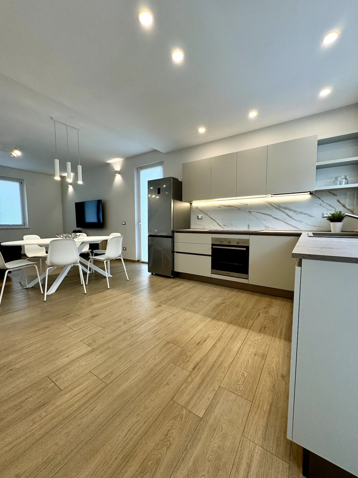 The modern kitchen features sleek cabinetry and a marble backsplash. A dining area includes a white table surrounded by six chairs. Soft lighting highlights the spacious layout, which contains a refrigerator and built-in oven integrated into the design.