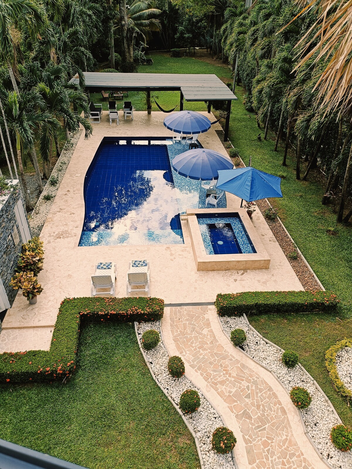 A large outdoor pool is surrounded by well-manicured grass and tropical landscaping. Two patio umbrellas provide shade over lounge chairs beside the pool. A hot tub is integrated into the pool area, with a stone pathway leading through vibrant flower beds.