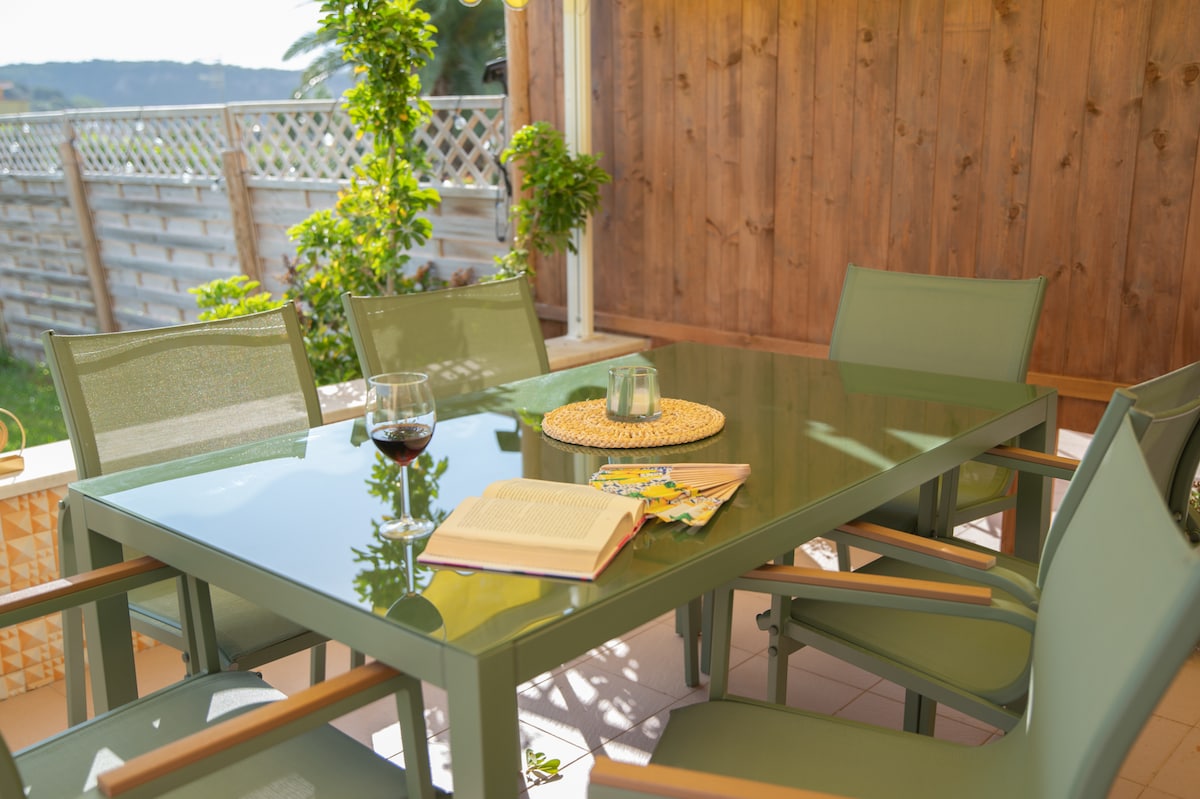 A dining table with a glass top is surrounded by several light green chairs. A book lies open on the table, accompanied by a decorative placemat and a glass of red wine. Lush greenery is visible in the background.
