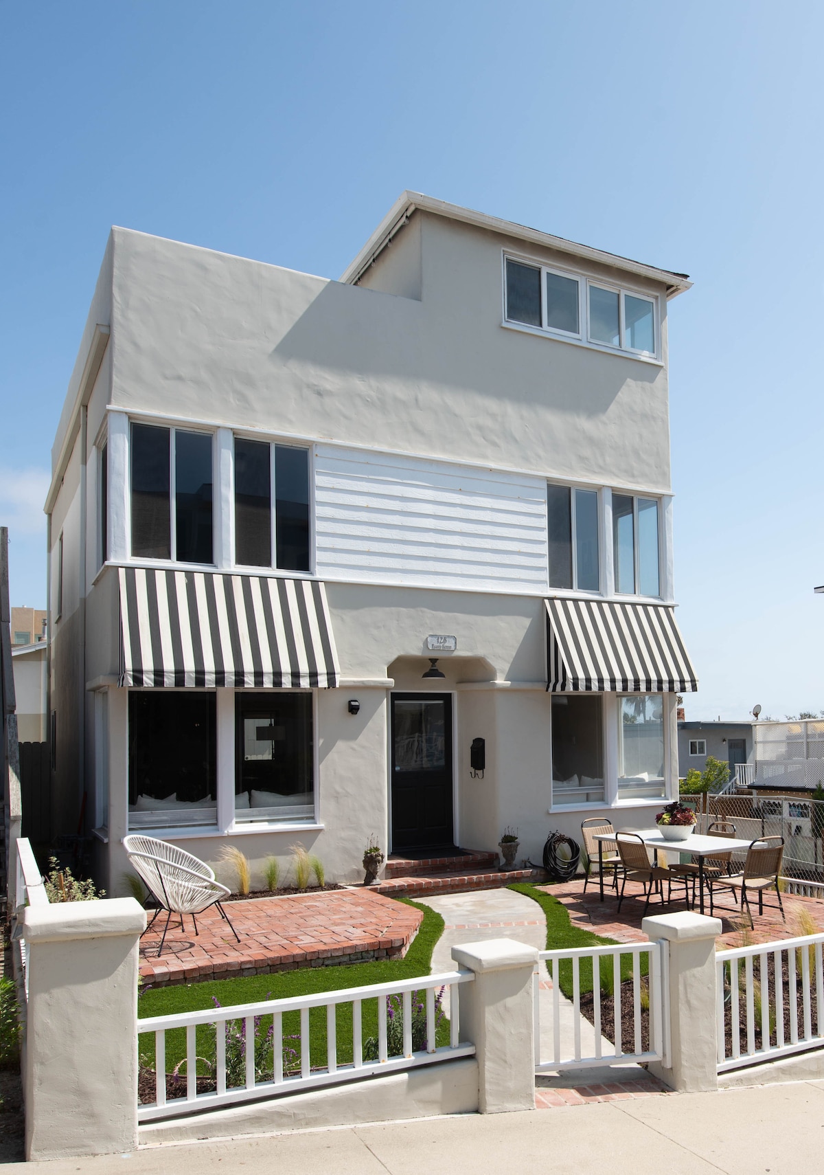 The exterior of a three-level beach house is shown, featuring large windows and striped awnings. A welcoming front patio includes a dining set among manicured grass. The house is painted in a light color, providing a modern ambiance.