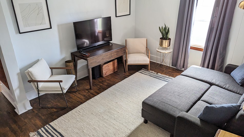 A living room area features a modern gray sectional sofa, complemented by a minimalistic white chair and a small round side table. A large television is positioned on a wooden console. Natural light filters in through a window, accentuating the warm wood flooring.