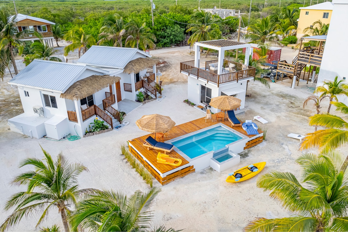 An aerial view of the property showcases several casitas surrounding a central pool area. The pool is framed by a wooden deck furnished with loungers and umbrellas. Nearby, a kayak rests on the sandy ground, with palm trees adding a tropical touch to the setting.