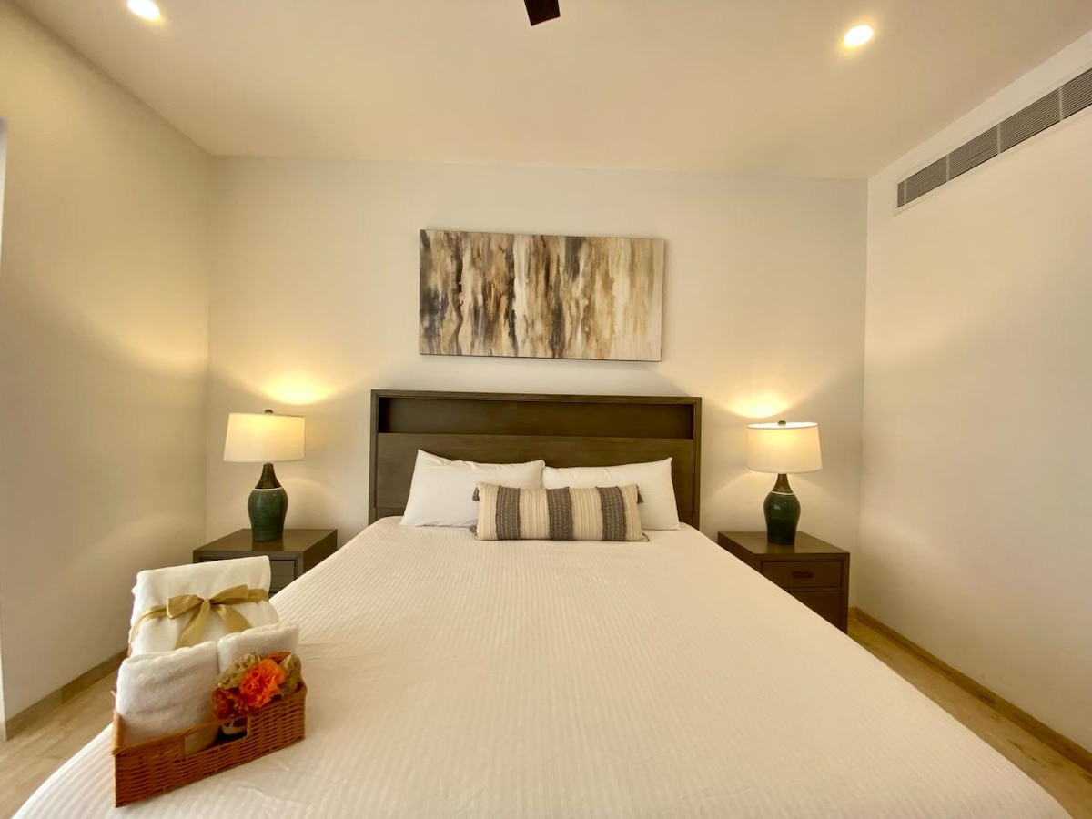 A spacious bedroom features a large bed with neutral bedding. Two bedside lamps provide soft illumination, and a piece of abstract artwork adorns the wall above the headboard. On the bed, a decorative tray holds folded towels and a small bouquet of flowers.